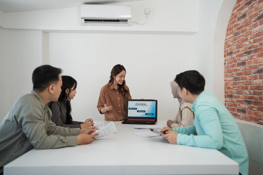 Young woman presents laptop slideshow to diverse coworkers during collaborative office workshop in bright modern room, with papers on table, attentive faces, and relaxed professional discussion.
