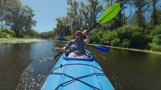 Mature woman paddling blue tandem kayak on calm river with green trees POV