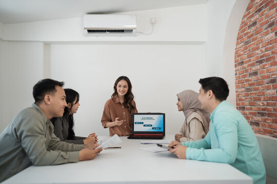 Diverse coworkers gather around table for collaborative office discussion with smiling presenter beside laptop screen and papers.