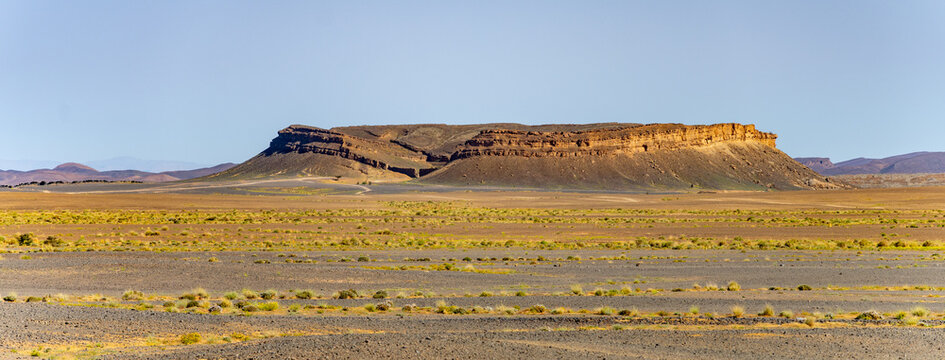 A wide view shows a rock formation in the dry desert of Morocco. The area is open with sparse vegetation and clear skies. The rock stands alone in the vast land.