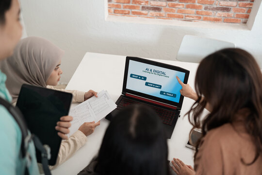 Students gather around laptop in bright modern classroom workspace, reviewing digital learning material, reading printed notes, and one person points at screen during discussion.