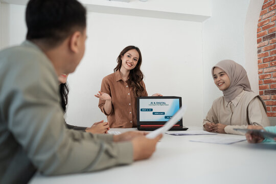 Smiling presenter leading office discussion with laptop screen visible while colleagues sit around conference table, reviewing documents in bright modern workspace