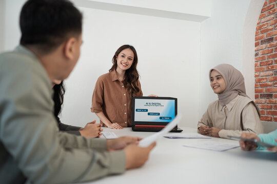 Smiling presenter stands next to open laptop as diverse teammates review paperwork during collaborative workshop in bright modern office with clean minimalist decor.