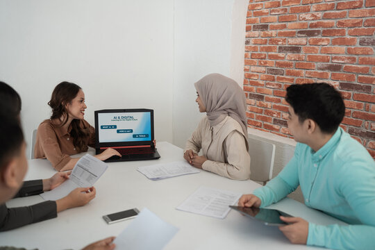 Smiling team members sit together reviewing printed documents and laptop screen in bright office with brick wall, showing collaboration, learning, and planning.