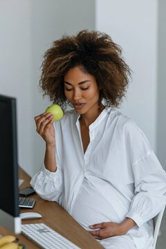 Pregnant woman at desk smelling a green apple for prenatal wellness background and healthy workplace banner design