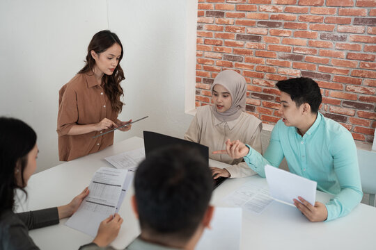 Young professionals review project documents on white desk during collaborative office meeting, with standing presenter using tablet, laptop, charts, and teamwork.