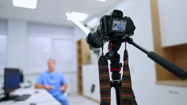 Tripod head and camera recording a seated healthcare presenter. A hanging strap and pan handle frame the camera display, with a man in blue scrubs blurred behind.