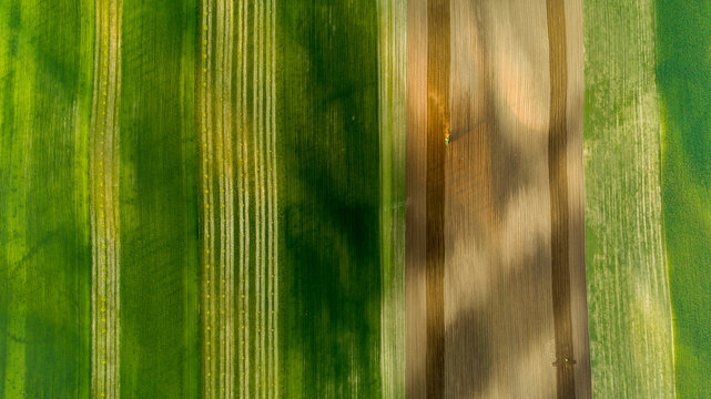 Aerial view of agricultural fields with alternating green and brown strips, showing a tractor working on the land under a bright sky in Brno-City District, South Moravian Region, Czechia.