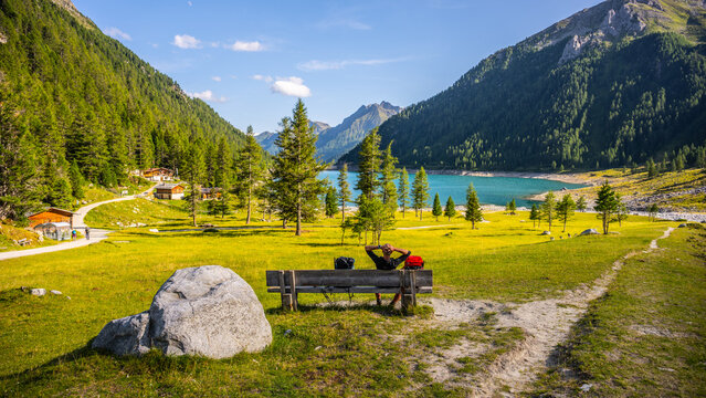Visitors relax by a bench at Lake Neves, located in South Tyrol at the end of the Molini Valley in the Zillertal Alps, Italy. The scene shows green fields and mountains surrounding the lake.