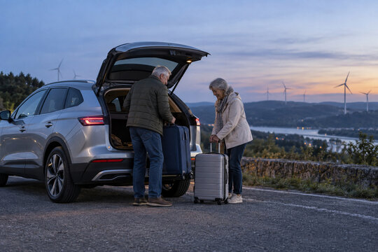 Senior couple organizing luggage beside a generic electric crossover at blue hour for regional road travel and active retirement campaigns