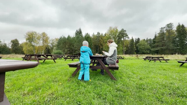 A child and mother rest comfortably at a wooden picnic table, sharing a snack. This cozy moment brings warm happiness in Pavlovsk Park, Saint Petersburg.