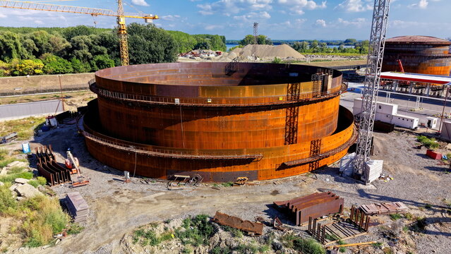 Aerial view of the assembly of a large industrial oil storage tank at a refinery construction site, featuring rusted steel panels, cranes, and heavy machinery in Kovin, Vojvodina, Serbia.