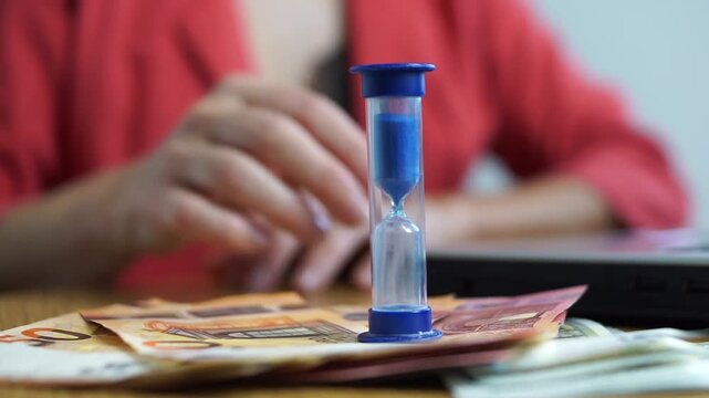 Businesswoman in red jacket placing a blue sand hourglass on top of euro banknotes with a laptop in the blurred background, financial planning and deadline concept