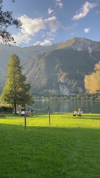 Lakeside park at sunset with calm lawn, slack volleyball net, and lifebuoy on wheeled cart near dock under tall leafy trees quiet recreation by shoreline glow