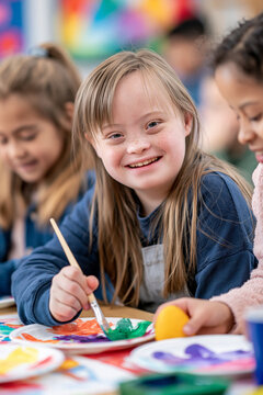 Blue sweatshirt on smiling girl with Down syndrome holding paintbrush next to black friend during art class, inclusive special education