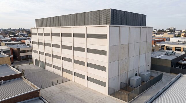 Multi Story Data Center Rising Among Low Urban Buildings In An Aerial View Of A Pale Concrete Tower With Grey Louvre Bands And A Rooftop Plant Screen On An Overcast Day