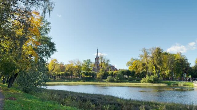 A historic church spire rises behind glowing autumn trees and a serene pond. Golden leaves bring peaceful joy to the calm water. This park creates tranquil harmony in Pushkin, Saint Petersburg.