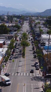 Aerial view shows vehicles driving along a sunny boulevard lined with tall palm trees, shops, and mountains visible in Los Angeles, California.