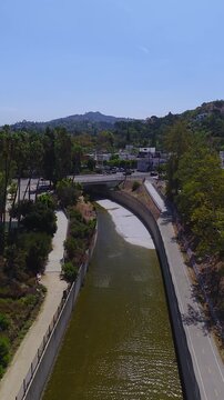 Aerial view following the concrete banks of an urban river channel in Los Angeles, flanked by bike paths, palm trees, and distant hillside neighborhoods on a sunny day.