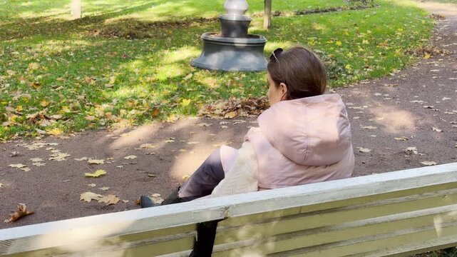 A woman sits peacefully on a wooden bench in a sunny autumn park. She rests quietly among fallen golden leaves. This serene moment was captured in Pushkin, Saint Petersburg.
