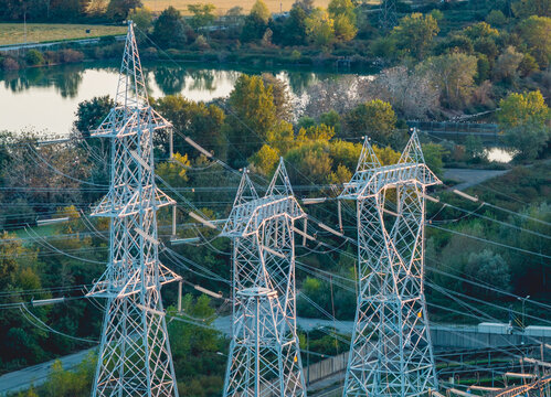 Aerial view of high voltage transmission pylons and power lines surrounded by lush vegetation near an industrial energy complex in Trino, Piedmont, Italy.