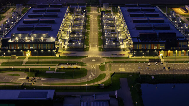 Aerial view of the Microsoft Data Center at night, featuring illuminated industrial buildings, parking lots, and a central access road in Middenmeer, North Holland, Netherlands.