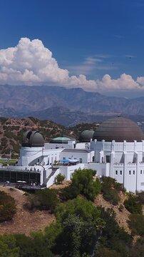 Aerial view of Griffith Observatory set against rolling hills, distant mountains, and clouds under a bright sky in Los Angeles, California, with visitors visible on the observation terraces.