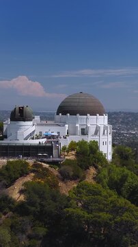 Aerial view of Griffith Observatory perched on Mount Hollywood in Los Angeles, California, with visitors walking on terraces surrounded by hillside vegetation under a bright sky.