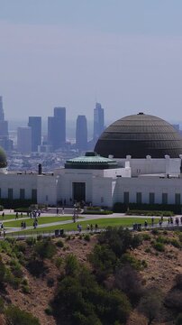 Aerial video showing Griffith Observatory on Mount Hollywood with tourists walking on grounds and the Los Angeles downtown cityscape visible across hazy horizon under daylight.