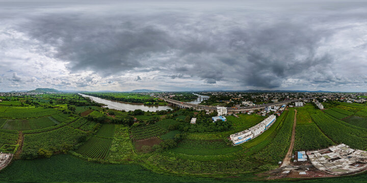 Aerial view of the Krishna River flowing through lush green agricultural fields and the cityscape of Karad with a bridge under a cloudy sky in Karad, Maharashtra, India.