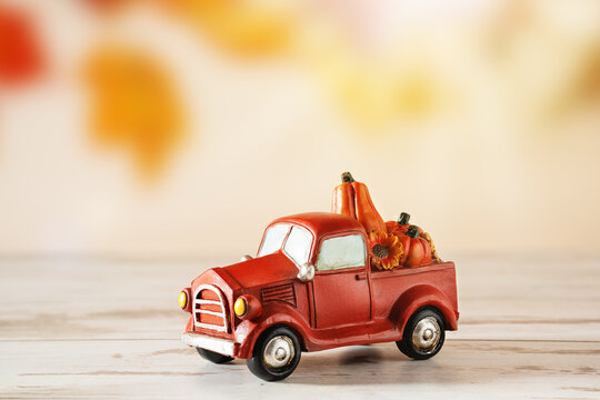 Toy red pickup truck figurine carrying pumpkins and sunflower on wooden table. Autumn harvest decoration with leaf blurred background and copy space for Thanksgiving and seasonal designs.