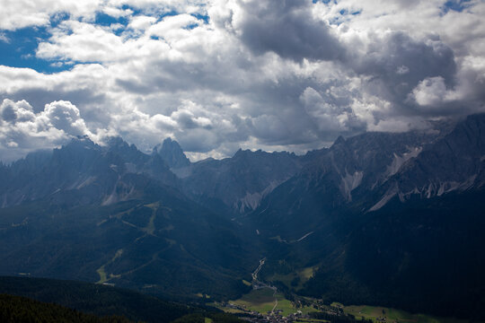 Aerial view of the dramatic Dolomites mountain range with rugged peaks, lush green valleys, and dense forests under a cloudy sky in Rocca Pietore, Veneto, Italy.