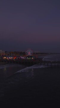 Aerial perspective over ocean waves reveals Santa Monica Pier glowing with amusement park rides, the iconic Ferris wheel, and night lights along the California coastline at twilight.