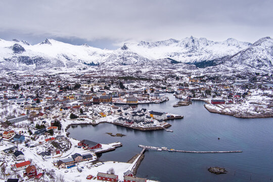 Aerial view of the snowy coastal town of Lofoten with colorful houses, a harbor, and dramatic snow-capped mountains under a cloudy sky in Nordland, Norway.