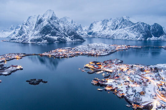 Aerial view of the snowy village of Lofoten with illuminated houses reflecting in the calm water, surrounded by dramatic snow-covered mountains in Nordland, Norway.
