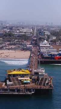 Aerial view of Santa Monica Pier stretching into the Pacific Ocean with people walking along the boardwalk, amusement park rides, sandy beach, end of Route 66 in Los Angeles, California.