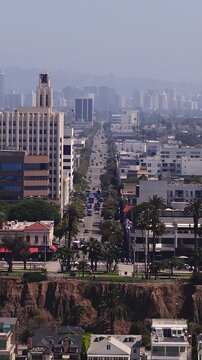 Aerial view shows traffic moving along a long street in Santa Monica past coastal cliffs, palm trees, and historic city architecture leading toward distant skyscrapers in California.
