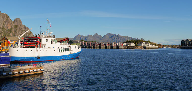 Sunny harbor scene in Svolvaer on the Lofoten Islands with large ship, traditional houses by the water, blue sea and impressive rocky mountains in the background.