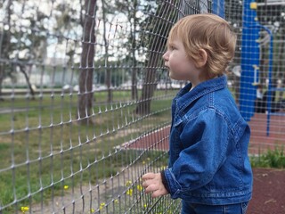 Toddler girl in denim jacket looking thoughtful near sports ground fence © Nastya