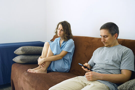 Sad woman sits hugging knees while partner uses smartphone, ignoring her presence. Portrays digital-addiction impact, phubbing behavior, loneliness within relationship, and lack of communication.