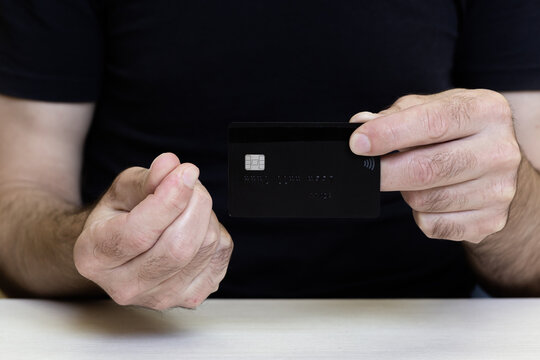 A man in a black T-shirt holds a credit card in front of him and makes a "money" gesture with his other hand.