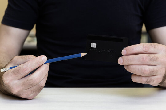 A man in a black T-shirt holds a black credit card in front of him and points to it with a blue pencil held in his other hand.