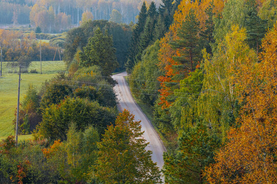 Winding rural road through colorful autumn forest and green fields beneath hazy wooded hills, tranquil countryside landscape viewed from an elevated perspective in fall