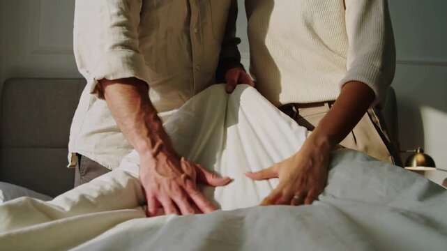 Lifting duvet peak, partners wearing button shirt and knit top smoothing bed in bedroom to tidy