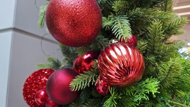 Shiny red glitter and glass baubles hang on green fir branches of a Christmas tree.