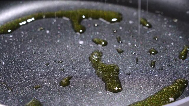 Close-up of black nonstick pan surface with scattered oil droplets, greenish highlights, glossy texture, plus rim under smooth kitchen light