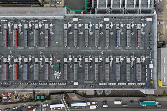 Aerial view of a large data center roof featuring rows of cooling units and ventilation systems, with a busy road and vehicles visible at the bottom edge, United Kingdom.