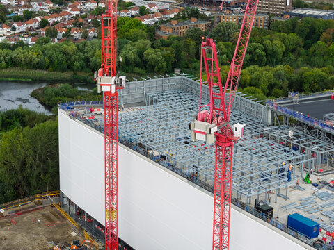 Aerial view of a data center construction site with two red tower cranes, exposed steel framework, and white wall panels surrounded by trees in the United Kingdom.