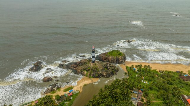 Aerial view of the iconic black and white striped Kapu Light House situated on a rocky outcrop surrounded by the Arabian Sea, sandy beach, and lush palm trees in Udupi, Karnataka, India.