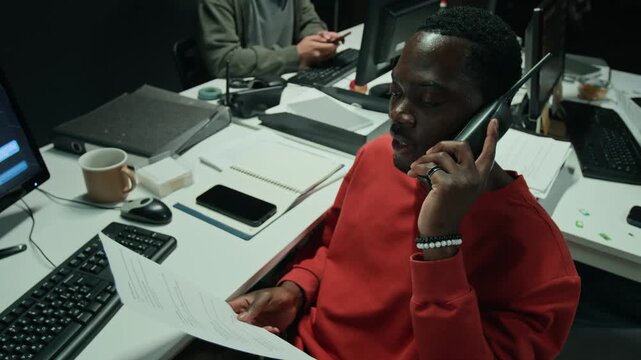 Young African American man talking on walkie talkie while checking scam documents near computer monitor. Clip suitable for cybersecurity projects and scam awareness
